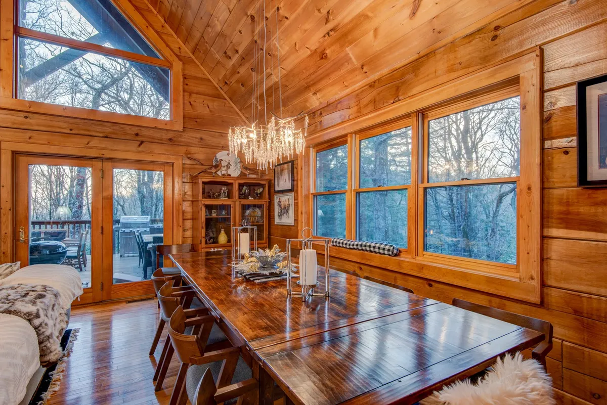 A large wooden dining table sits beneath a crystal chandelier, framed by expansive windows with breathtaking nature views. The setting is both intimate and grand, ready for family feasts or special celebrations.