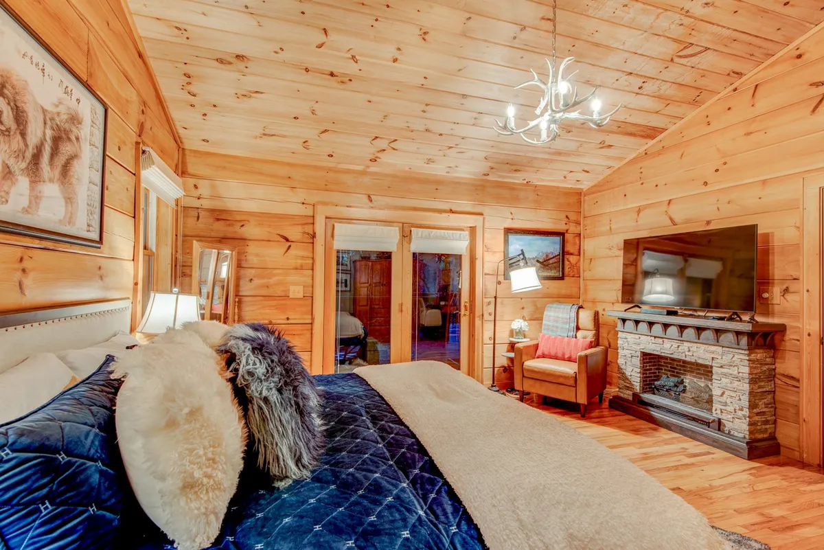 A warmly lit bedroom featuring a plush bed with soft pillows, a prominent stone fireplace, a wooden ceiling, tasteful artwork on the walls, and a mounted television. The room invites relaxation with cozy blankets, and the glow from the fire creates an inviting ambience.