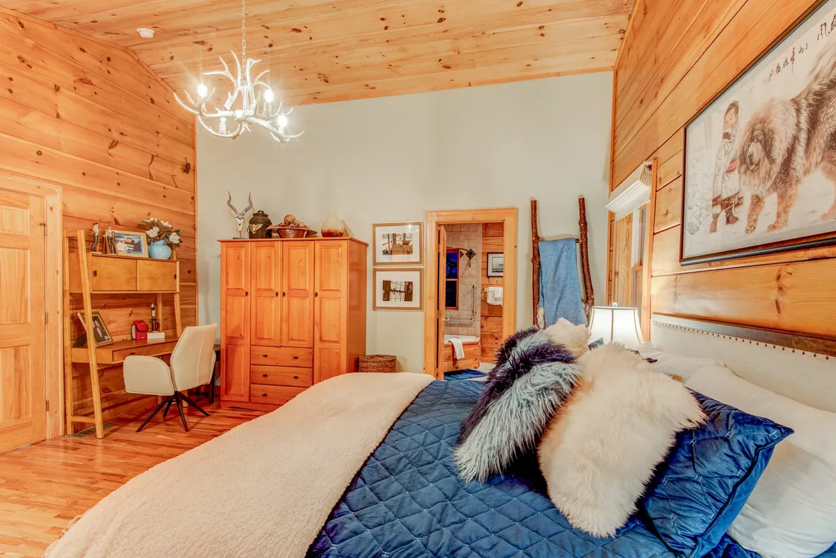 A serene bedroom with a plush bed layered in soft linens, wooden furnishings, a chandelier light fixture, and decorative artwork. Gentle natural light filters into the room, evoking a warm and restful atmosphere ideal for a quiet morning awakening.