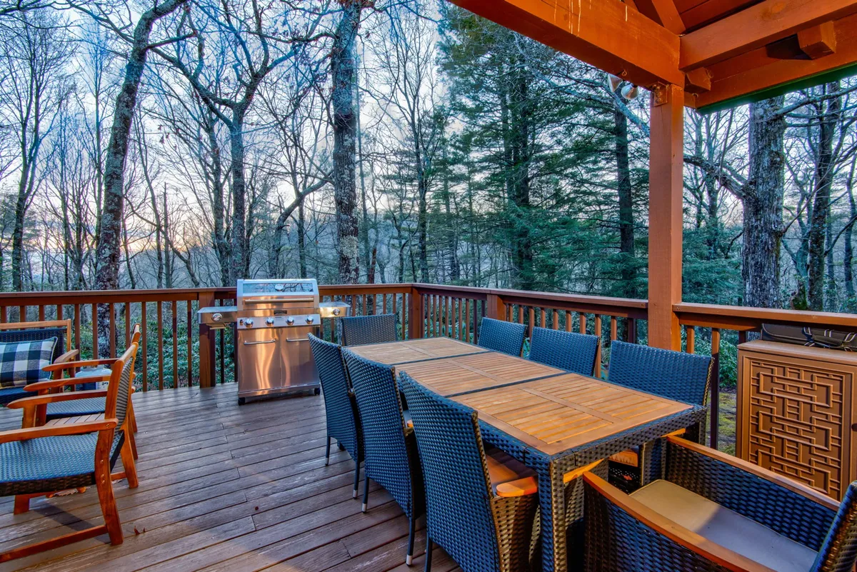 An outdoor dining area featuring a large wooden table, an outdoor grill, woven chairs, and forest views. The space is perfect for al fresco meals, socializing, and enjoying the sights and sounds of nature.
