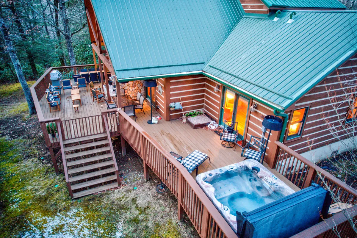 A cozy outdoor deck features an inviting hot tub, comfortable seating, wooden steps, and the glow of warm light from large windows. The natural surroundings hint at tranquil forest views, while the setup makes it ideal for relaxing in the hot tub, gathering with family, and stargazing under the open sky.