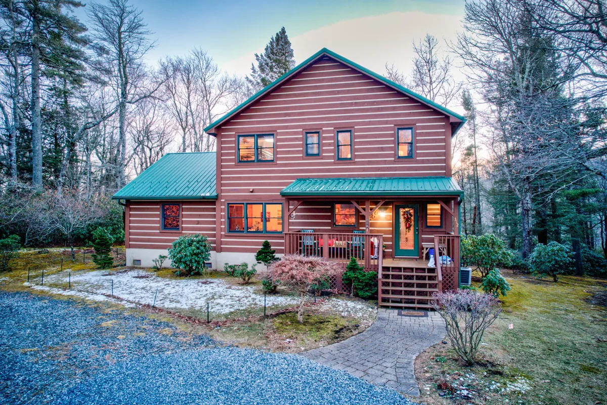 A classic log cabin structure with a green metal roof and a spacious wooden deck, set among lush greenery. The porch offers a welcoming space for relaxing outdoors, with views of the surrounding trees and natural landscape.
