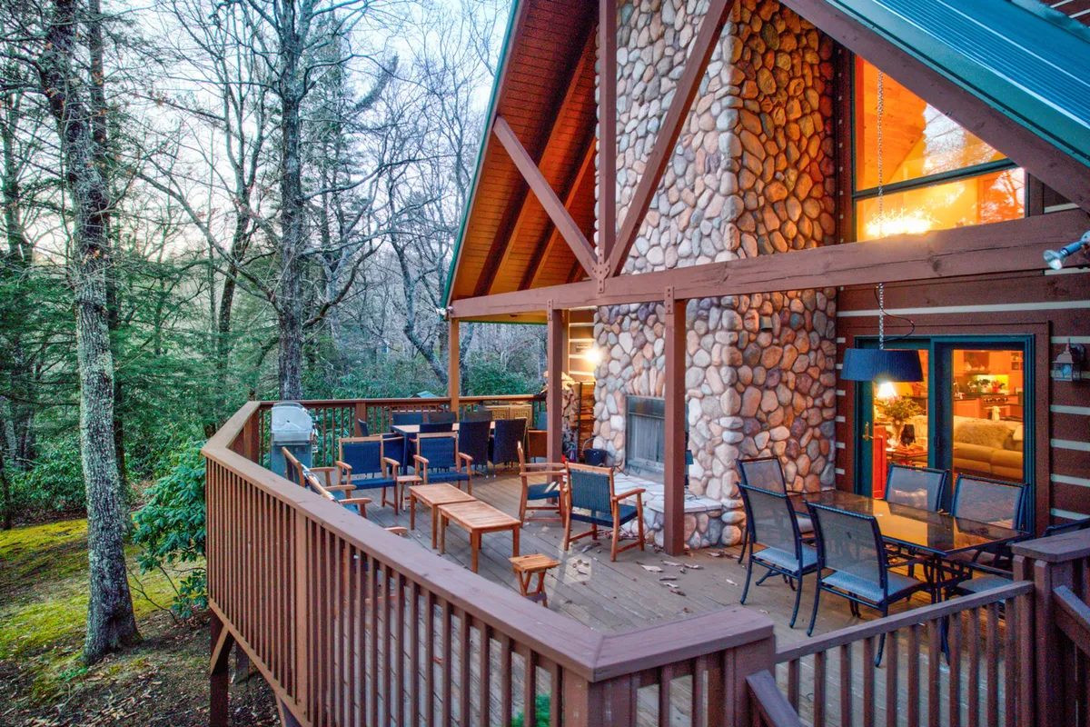 A large stone fireplace is the focal point of a spacious wooden deck, set against a backdrop of lush trees and large windows that reflect the outdoors. The space is designed for gatherings, with plenty of seating for guests to relax and enjoy the warmth of the fire.