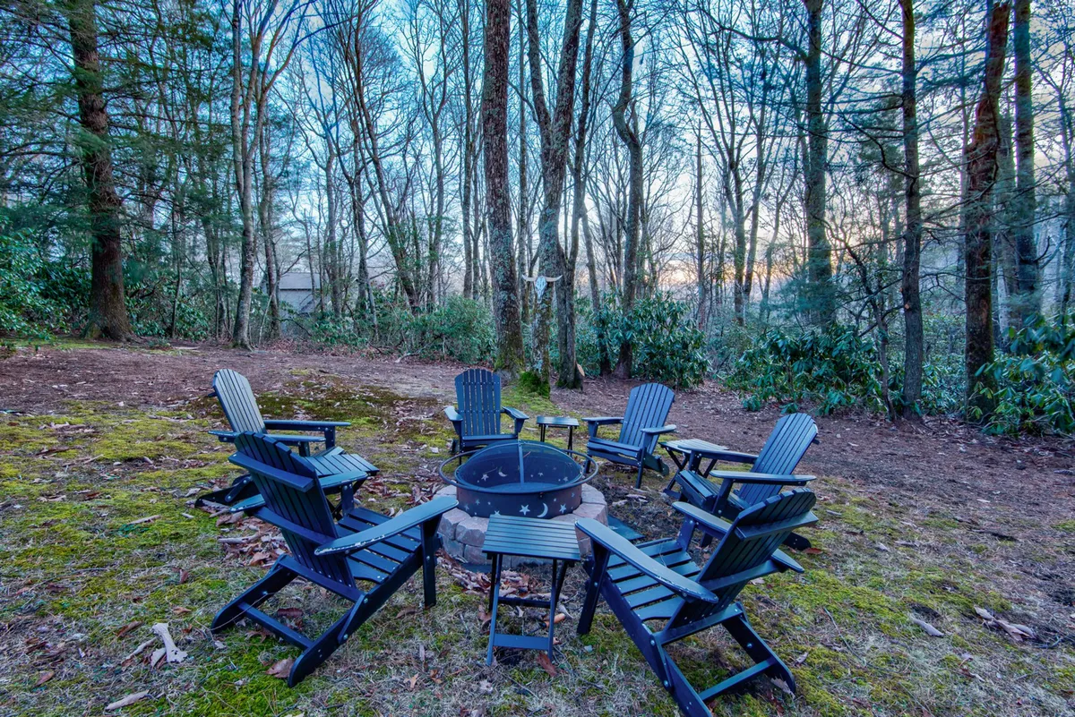 An inviting fire pit area with Adirondack chairs arranged in a circle, surrounded by tall trees. The scene suggests a relaxed, social atmosphere perfect for storytelling, roasting marshmallows, or stargazing on clear nights.