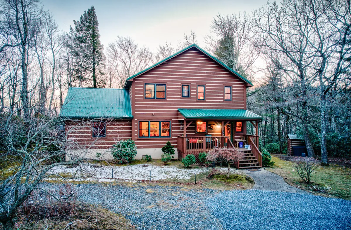 The image showcases a charming log cabin with a green metal roof, nestled among tall trees. A spacious front porch with a rustic railing invites guests to relax while soaking in the peaceful mountain surroundings. The overall scene conveys warmth, coziness, and a perfect blend of nature and comfort.