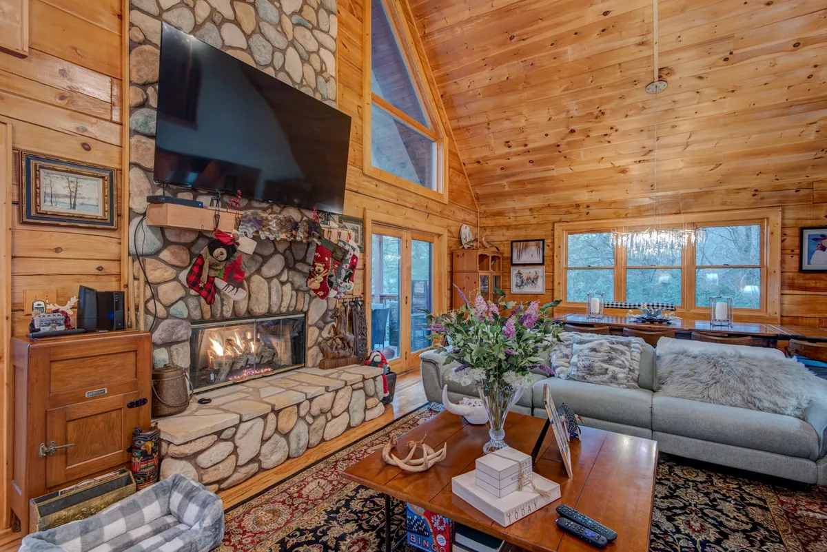 This image showcases a warm, welcoming great room with a striking stone fireplace as the focal point, flanked by expansive floor-to-ceiling windows that reveal beautiful forest views. A comfortable sofa sits in front of the fire, inviting gatherings and relaxation, while a floral arrangement adds a touch of homeliness.