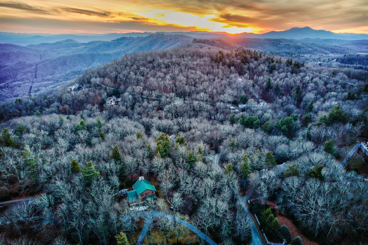 A panoramic view shows the wooden cabin surrounded by dense forest, with a stunning mountain range and sunset horizon in the background. The scene is tranquil and frames the property within the vast, beautiful landscape—perfect for hiking, nature walks, or simply admiring the changing seasons.