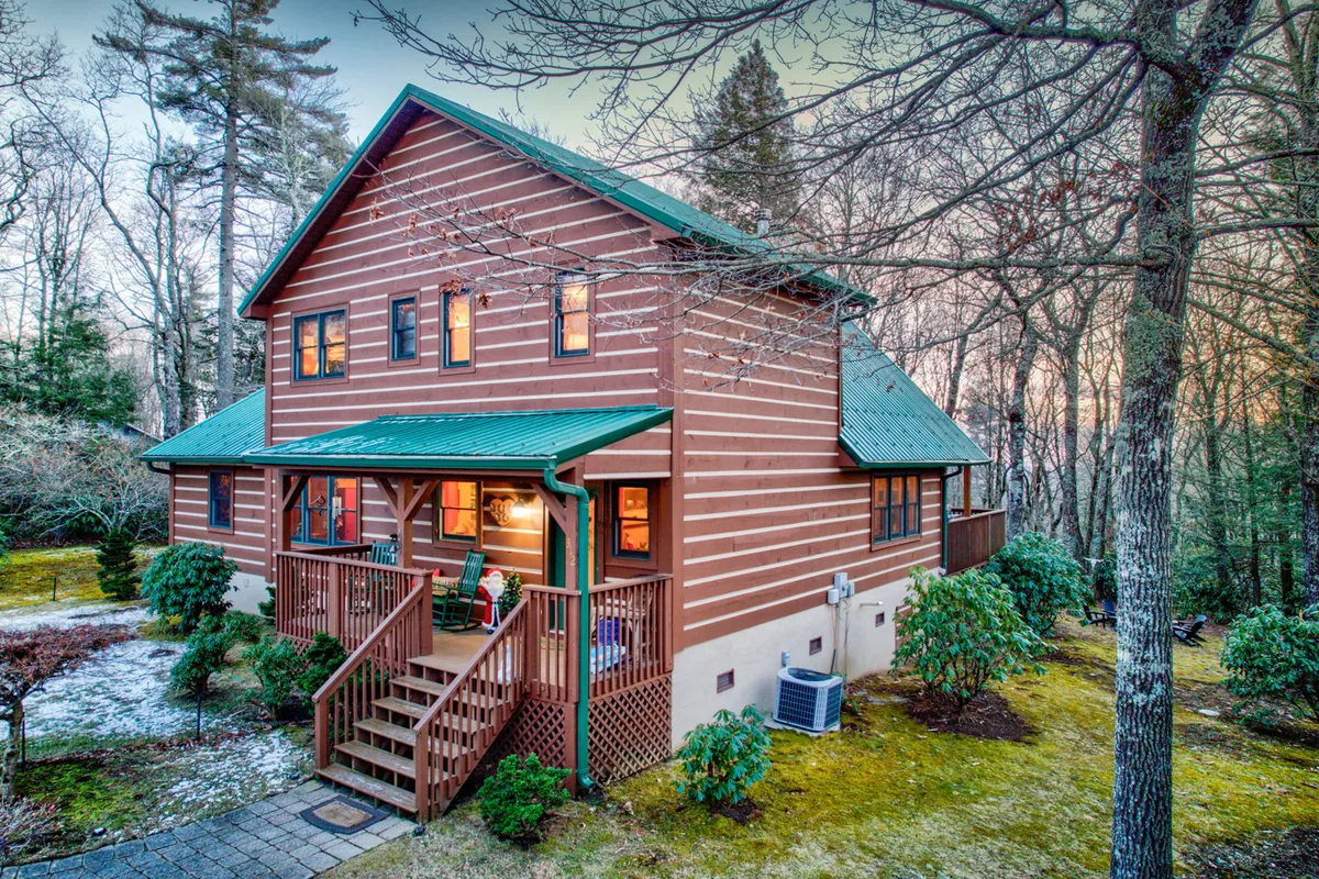 A classic wooden cabin with a green metal roof sits nestled among tall trees. The front porch is visible and evokes thoughts of outdoor gatherings, porch sittings, and nature walks. The surrounding foliage speaks to the seasonal beauty described in the section, from lush greens to vibrant autumn leaves or winter snow.