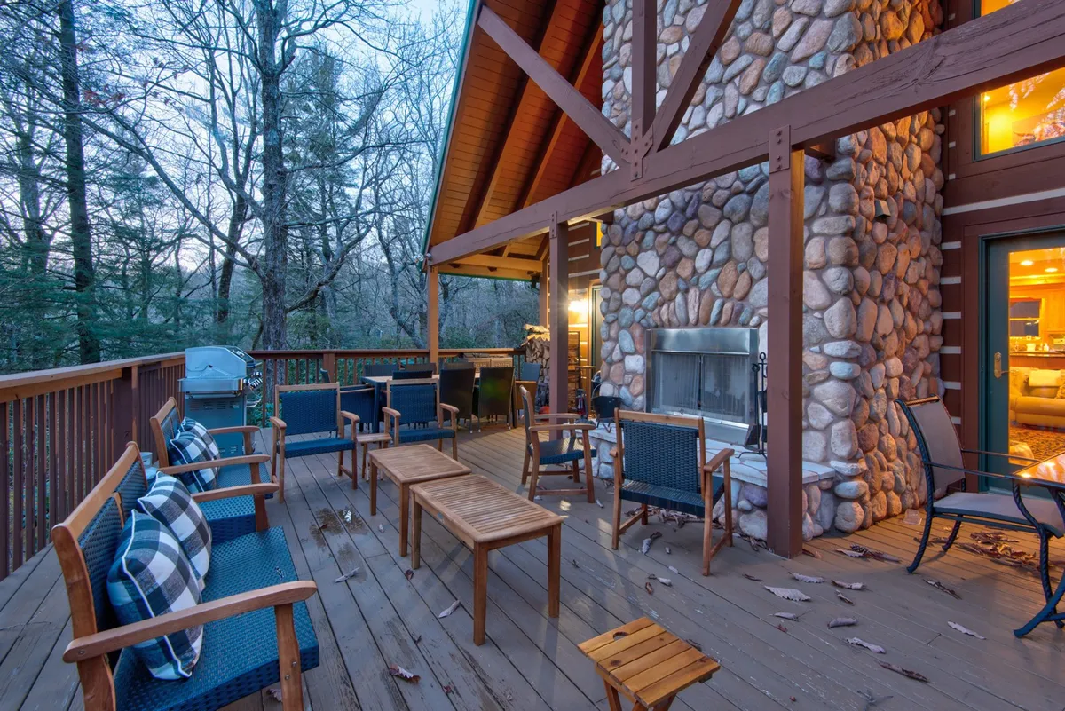 An inviting outdoor living area featuring a stone fireplace, spacious wooden deck, plush seating, and a grill. Surrounded by tall trees, this space exudes warmth and is perfect for evening gatherings, storytelling by the fire, or stargazing with friends and loved ones.