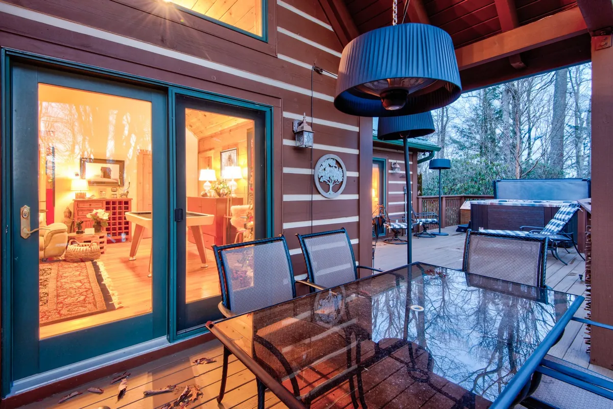 An exterior deck set for entertaining, with a large dining table, ambient hanging light, and a view into the warmly lit interior. The space hints at a hot tub nearby and provides an inviting atmosphere for outdoor meals and celebrations.