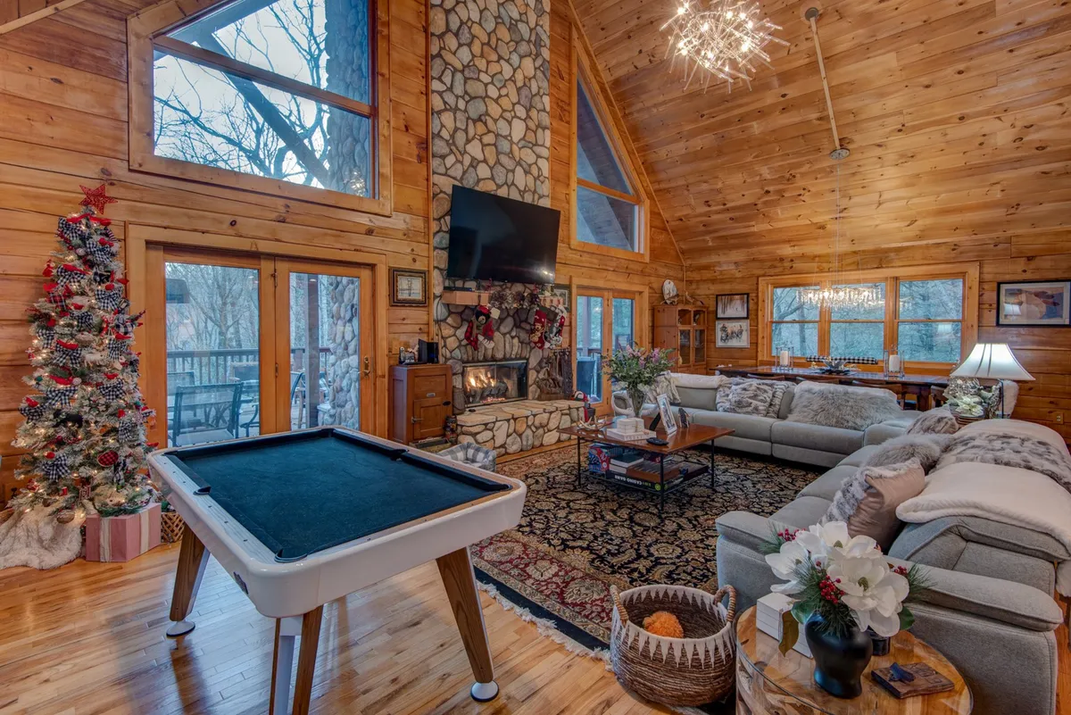 A vibrant interior focused on a pool table beside a fireplace, with a Christmas tree adding a festive touch and large windows letting in natural light. A comfortable sofa provides additional gathering space.
