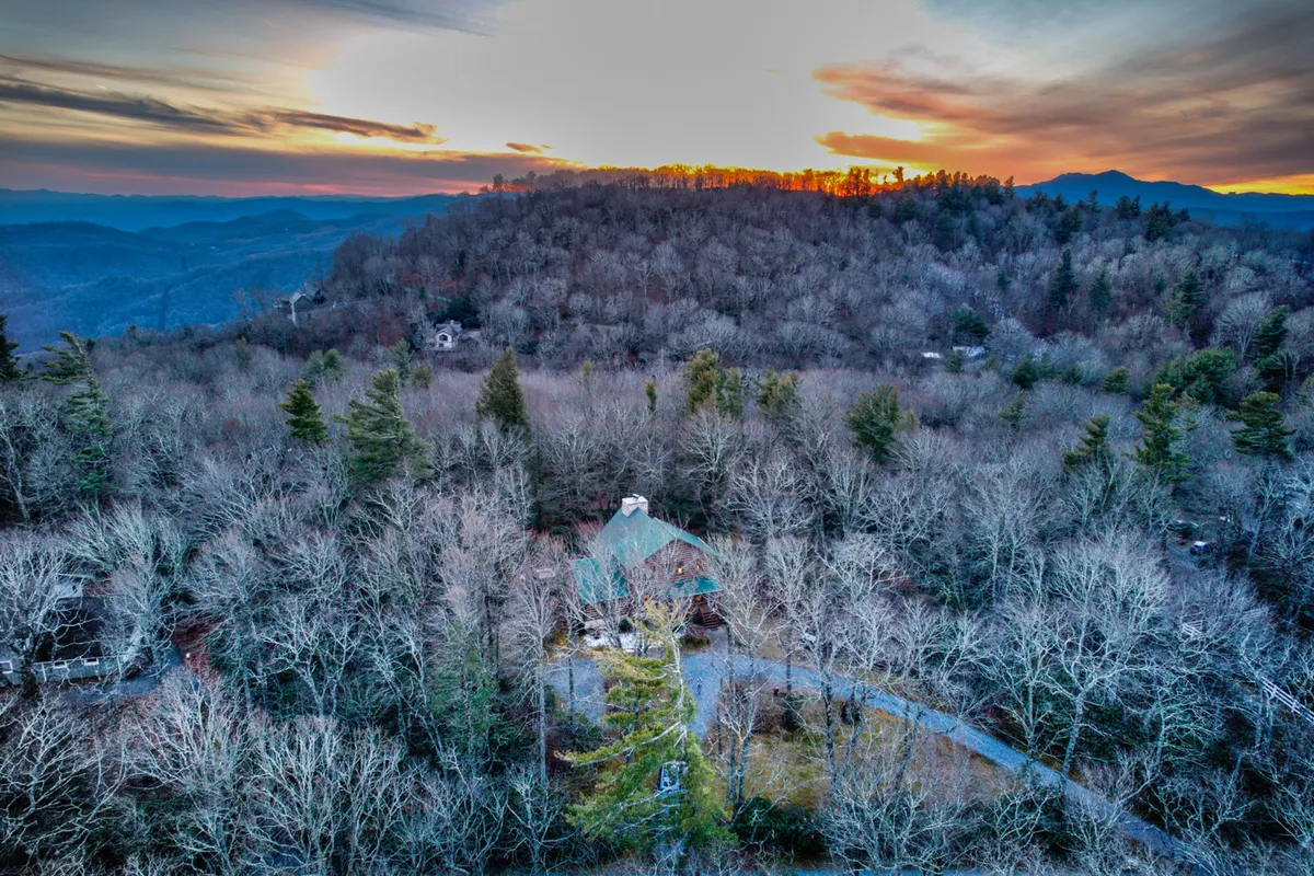 A stunning view of the house nestled among trees with distant mountains in the background, all beneath a vibrant sunset sky. The scene evokes tranquility, natural beauty, and the promise of adventure in the surrounding wilderness.